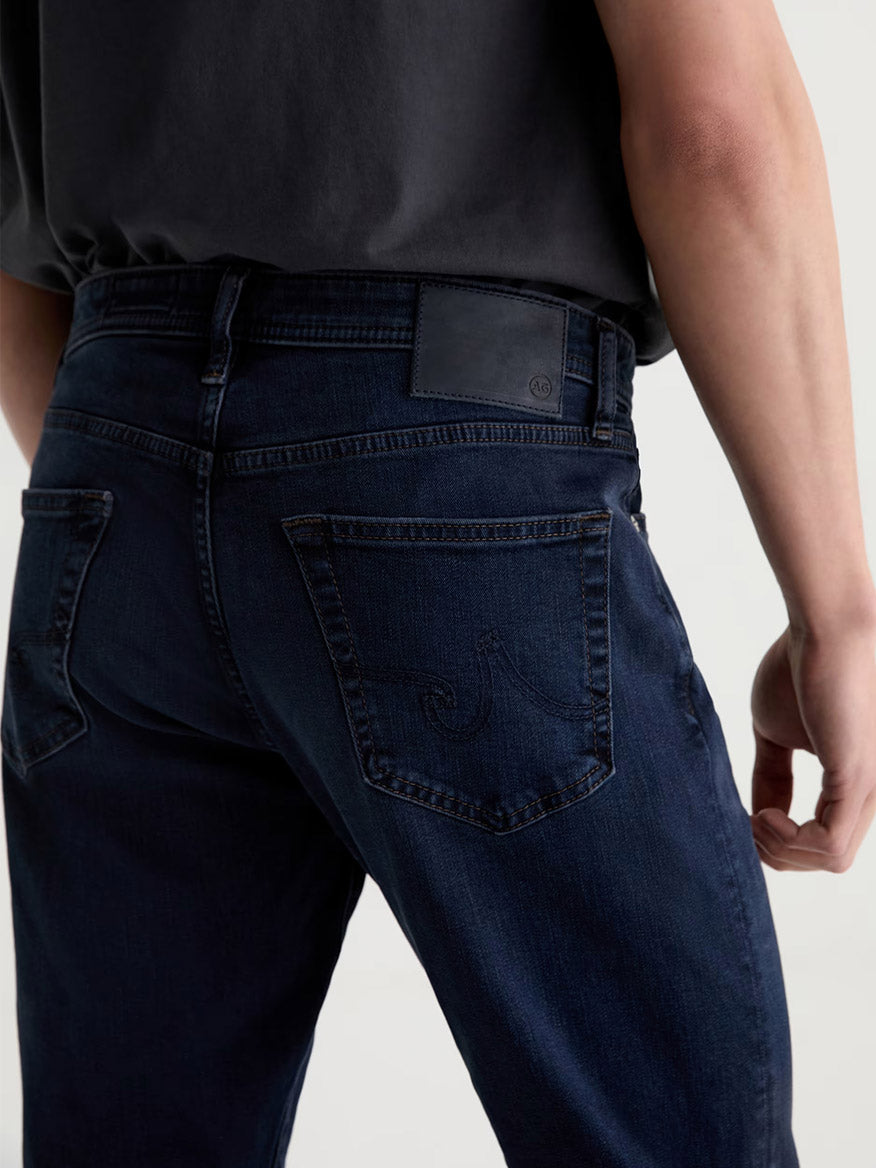 Person wearing the AG Jeans Graduate in Studio dark blue jeans and a black shirt, shown from the back at waist level, with visible back pockets and a black waistband label.