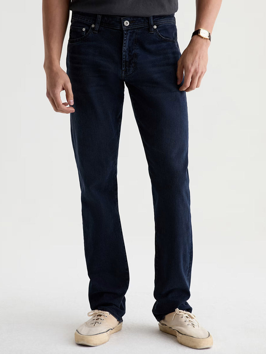 A person wearing the AG Jeans Graduate in Studio—dark blue straight-leg denim jeans—paired with a black t-shirt, white sneakers, and a watch, stands against a plain light background.