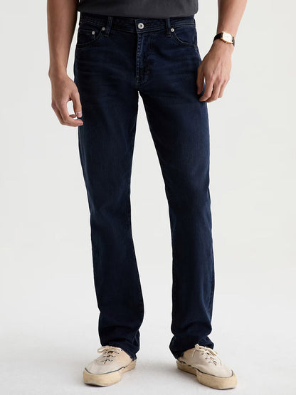 A person wearing the AG Jeans Graduate in Studio—dark blue straight-leg denim jeans—paired with a black t-shirt, white sneakers, and a watch, stands against a plain light background.