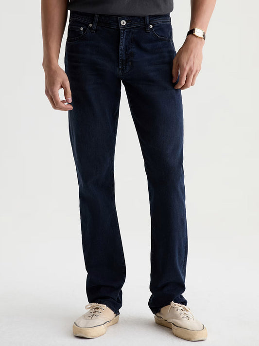 A person wearing the AG Jeans Graduate in Studio—dark blue straight-leg denim jeans—paired with a black t-shirt, white sneakers, and a watch, stands against a plain light background.