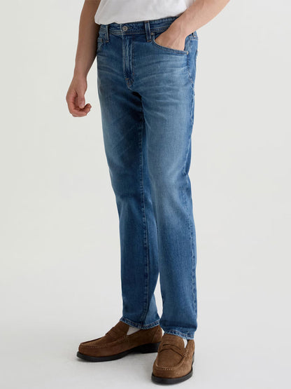 A person wears AG Jeans Graduate in Ashfield—tailored-leg premium denim—with a white shirt and brown loafers, standing against a plain light background, one hand in their pocket.