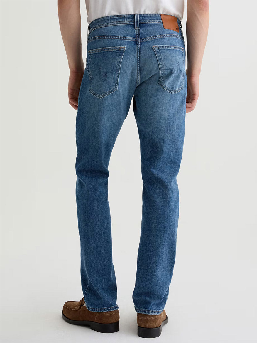 Someone is standing with their back to the camera against a plain background, wearing AG Jeans Graduate in Ashfield (premium denim blue jeans), brown shoes, and a white shirt.