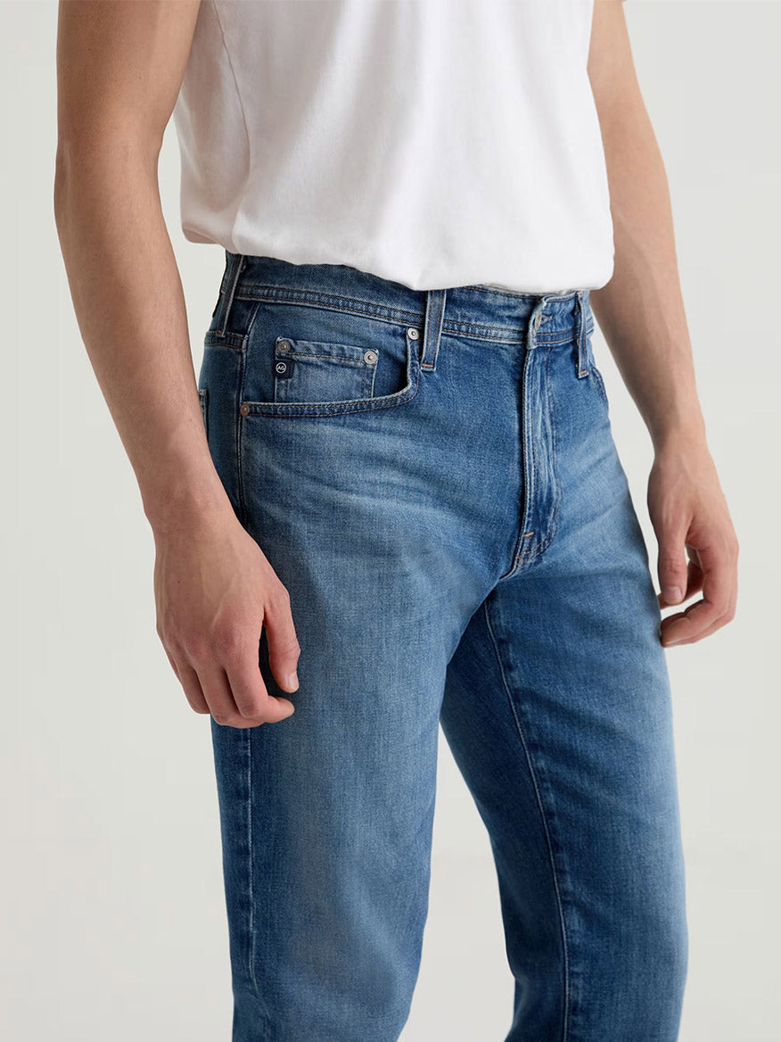 A person wearing a plain white T-shirt and AG Jeans Graduate in Ashfield, made from comfort stretch denim, stands against a neutral background. Only the torso and upper legs are visible.