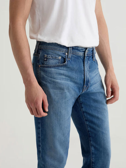 A person wearing a plain white T-shirt and AG Jeans Graduate in Ashfield, made from comfort stretch denim, stands against a neutral background. Only the torso and upper legs are visible.