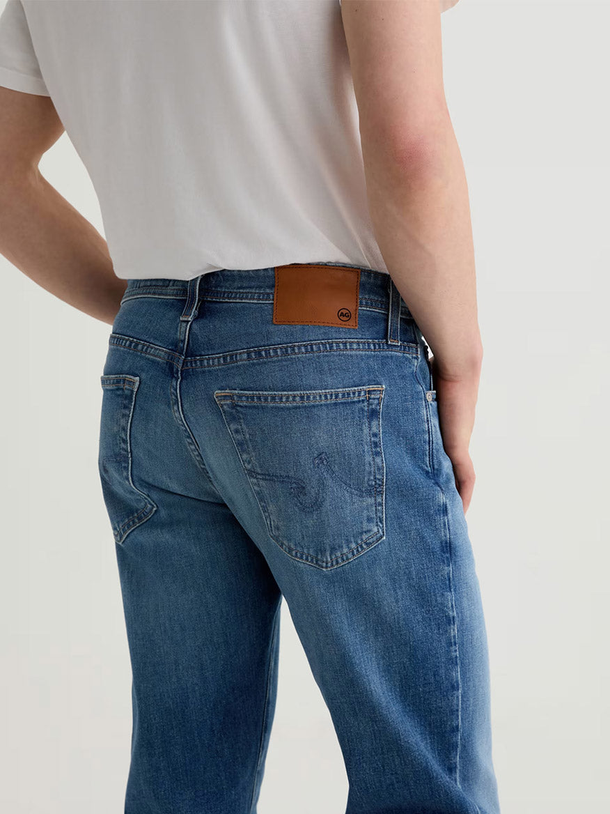 Model wearing AG Jeans Graduate in Ashfield, a light blue premium denim with men's tailored-leg fit and a brown leather patch on the waistband, paired with a white T-shirt, shown from the back.