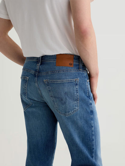 Model wearing AG Jeans Graduate in Ashfield, a light blue premium denim with men's tailored-leg fit and a brown leather patch on the waistband, paired with a white T-shirt, shown from the back.