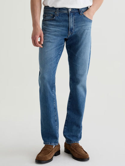 A person wears AG Jeans Graduate in Ashfield—men’s tailored-leg premium denim jeans—paired with a white shirt and brown suede shoes, standing against a plain light background.