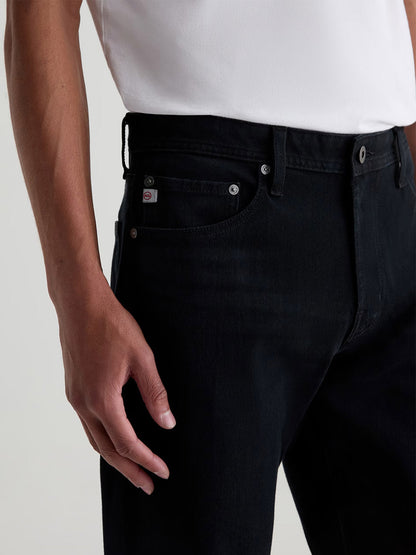 A person wearing a white shirt and AG Jeans Graduate in Vintage Ink is shown from the waist down, with one hand resting by their side.