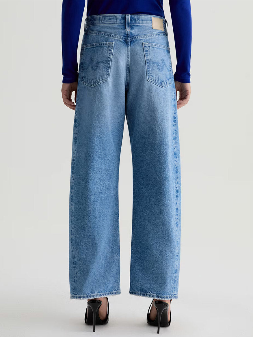 Person wearing AG Jeans Hattie High Rise Barrel in Blueprint, styled with black high heels, photographed from the back against a plain background.