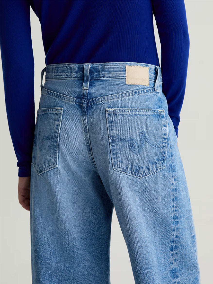 Person wearing AG Jeans Hattie High Rise Barrel in Blueprint, made from quality Sioux Rigid Denim by Candiani Mill. Shown from the back, these light blue jeans feature embroidered back pockets and a blank waistband patch.