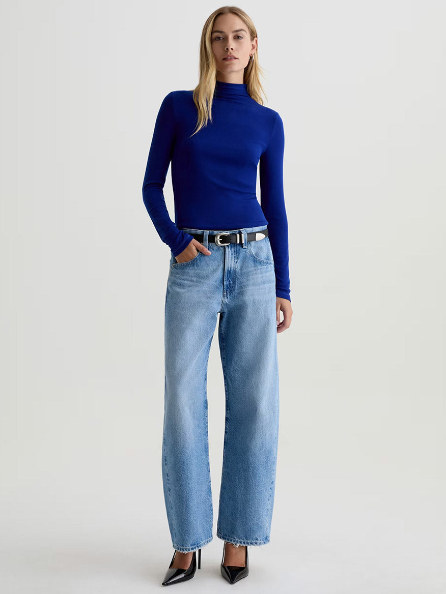 A person wearing a fitted long-sleeve blue turtleneck, AG Jeans Hattie High Rise Barrel in Blueprint, a black belt, and black pointed-toe heels stands against a plain background.