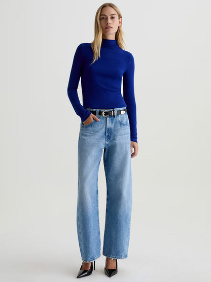A person wearing a fitted long-sleeve blue turtleneck, AG Jeans Hattie High Rise Barrel in Blueprint, a black belt, and black pointed-toe heels stands against a plain background.
