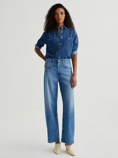 A woman faces forward, wearing the AG Jeans Hattie High Rise Barrel in Flashback with a denim shirt tucked in and cream shoes.