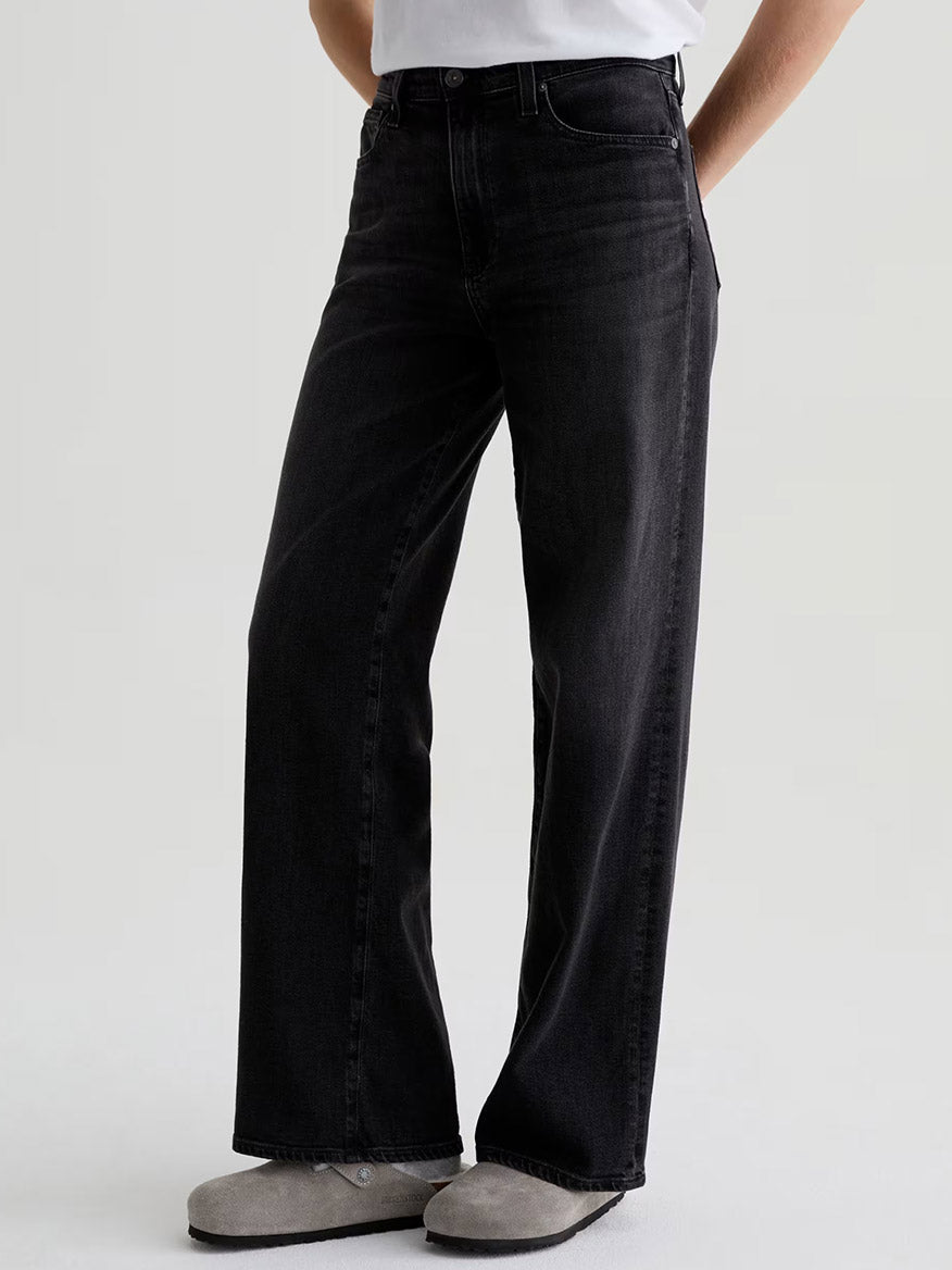 Model wears AG Jeans Kora High-Rise Wide Leg in Nebula, crafted from comfort-stretch denim, styled with a white T-shirt and gray slippers against a plain light background.