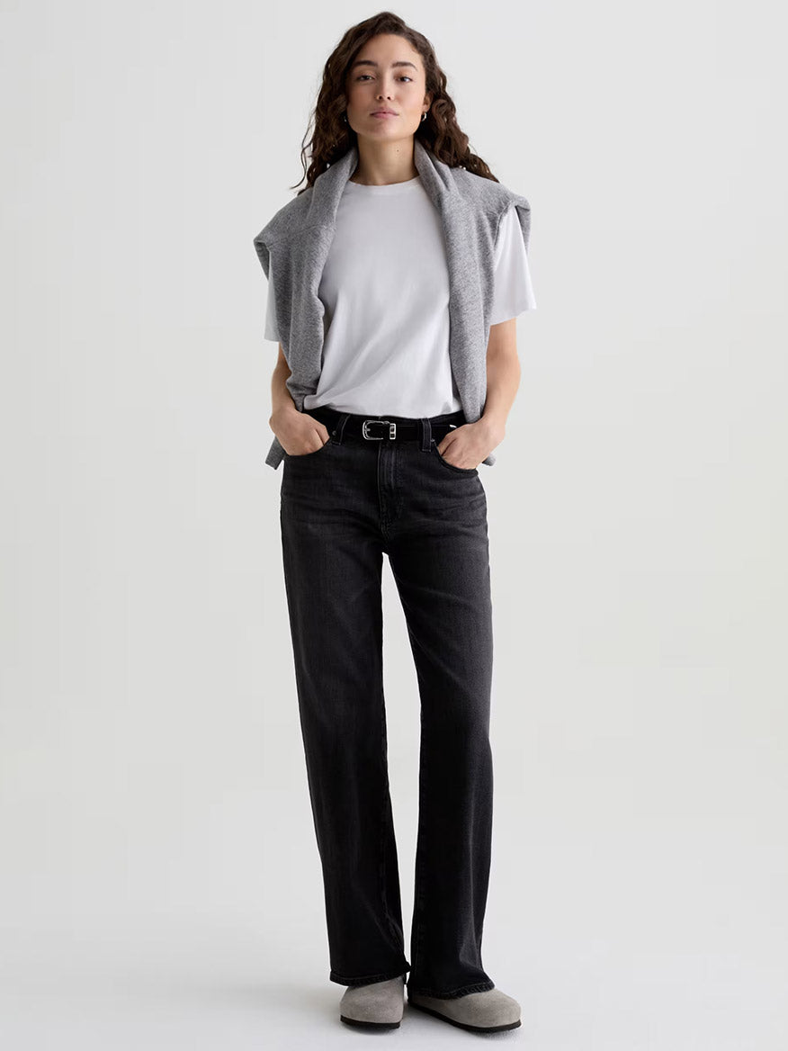 Person standing against a plain background wearing AG Jeans Kora High-Rise Wide Leg in Nebula, a white t-shirt, gray sweater draped over shoulders, and gray shoes. Hands are in pockets.