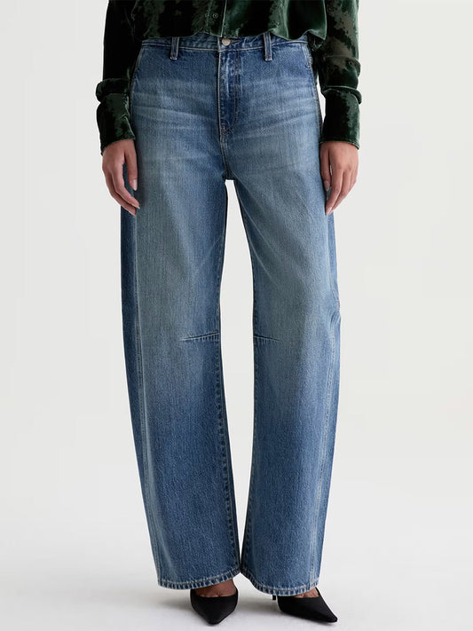 Wearing the AG Jeans Mercer Cuffed High-Rise Barrel Jean in Vibrato, paired with a dark green long-sleeve shirt and black pointed-toe shoes, this person stands against a plain background.