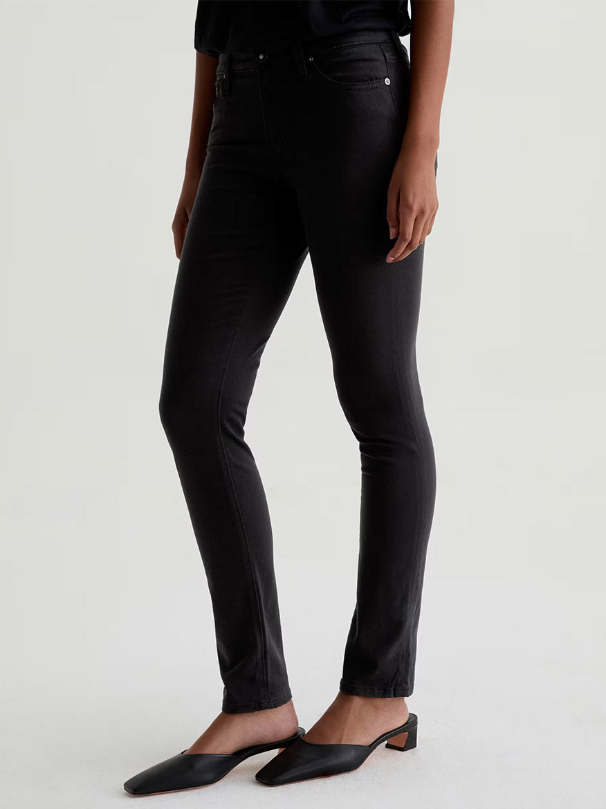 A person wears AG Jeans Prima Cigarette Leg in Super Black styled as skinny jeans with black low-heeled mules, standing against a plain light background.