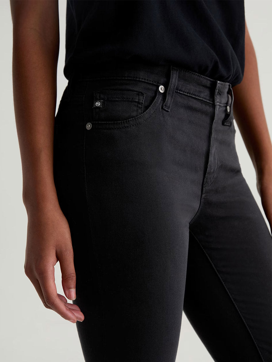 A person wearing AG Jeans Prima Cigarette Leg in Super Black and a black shirt, with their left arm relaxed at their side, shown from mid-torso to mid-thigh against a plain background.