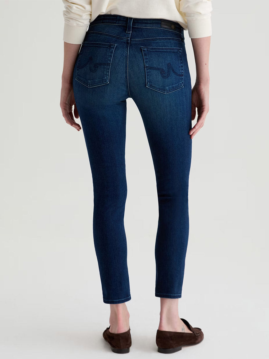Model wears the AG Jeans Prima Cigarette Leg in Epilogue, a dark blue ankle-length comfort-stretch denim jean, paired with brown flats. Photographed from the back against a plain light background.