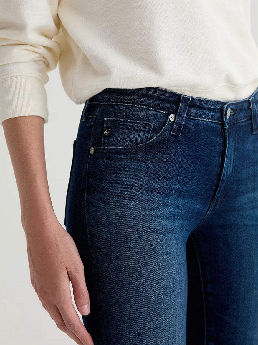 A person wearing a cream long-sleeve shirt and AG Jeans Prima Cigarette Leg in Epilogue, shown from mid-torso to upper thighs, with one hand relaxed at their side.