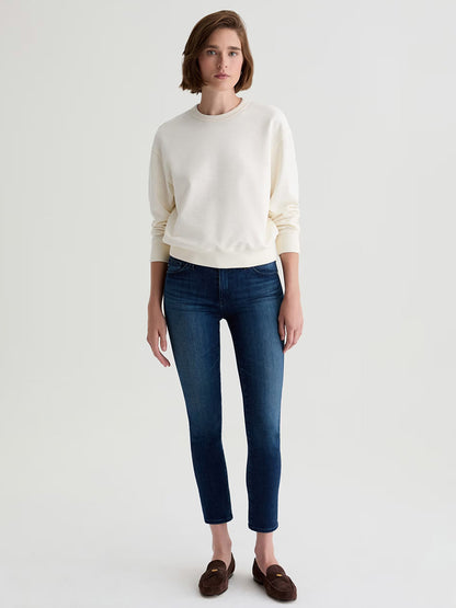 A woman faces forward wearing a white sweatshirt, AG Jeans Prima Cigarette Leg in Epilogue—ankle-length slim comfort-stretch denim—and brown loafers against a plain light background.