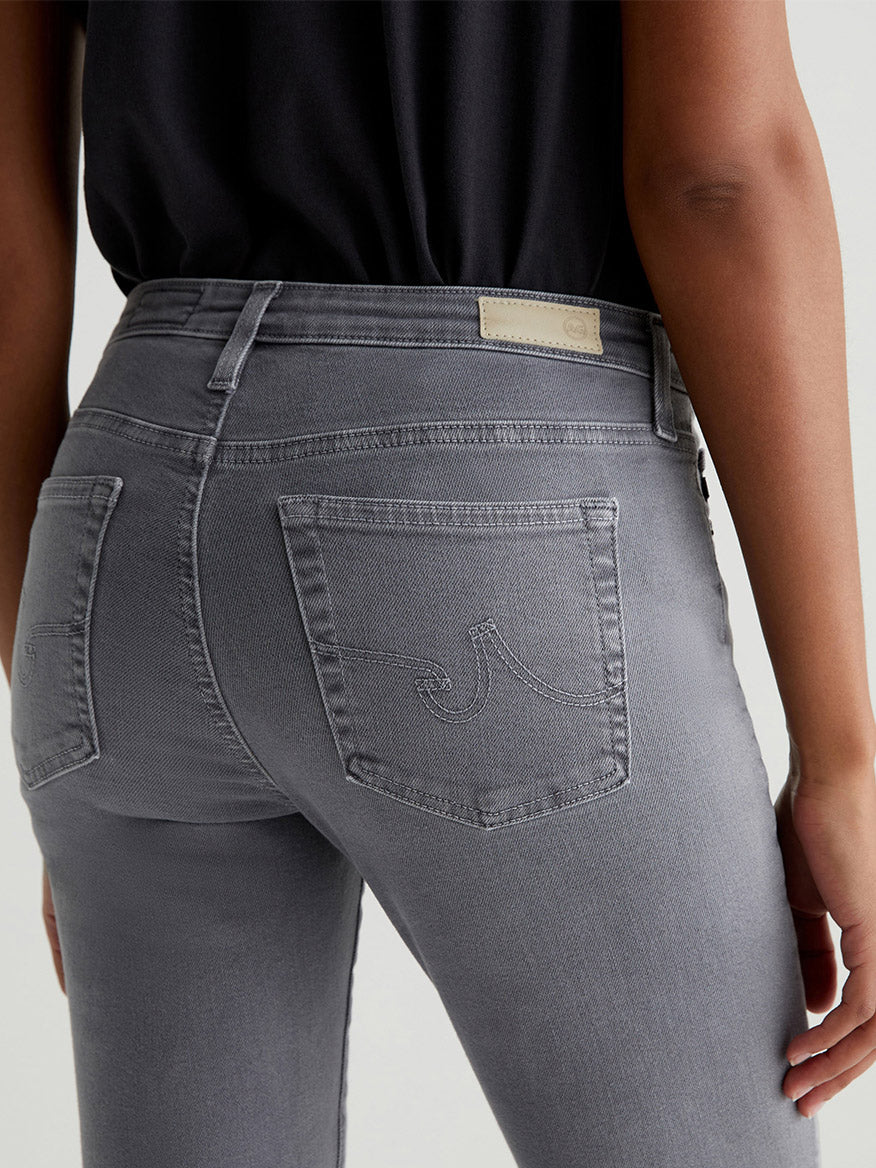 A person is shown from the back wearing AG Jeans Prima Cigarette Leg in Sulfur Grey Ember and a black top. These jeans, featuring a faded grey shade with two back pockets adorned with stitched designs, are crafted from Cloud Soft Stretch Denim for comfort.