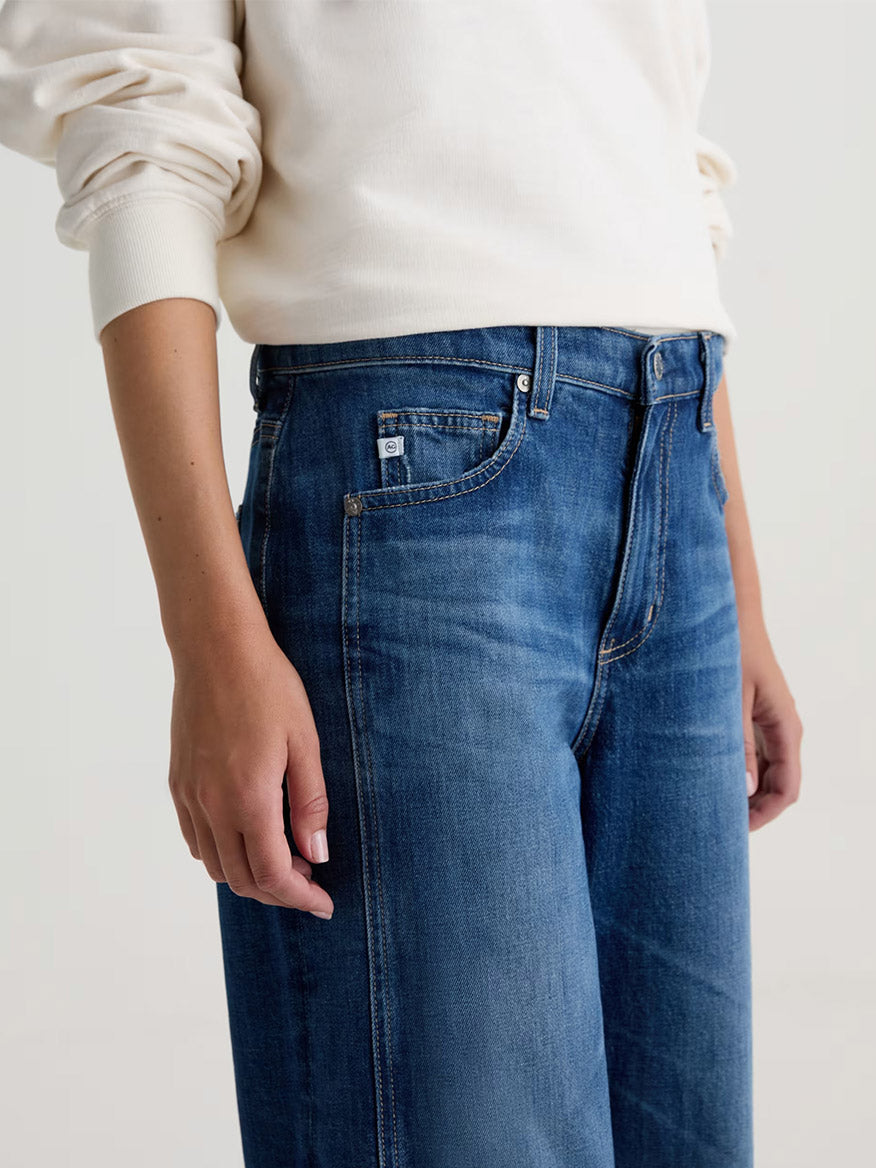 A person wearing a white long-sleeve shirt and AG Jeans Saige Wide Leg Crop in Watermist, shown from shoulders to mid-thigh, stands against a plain background.