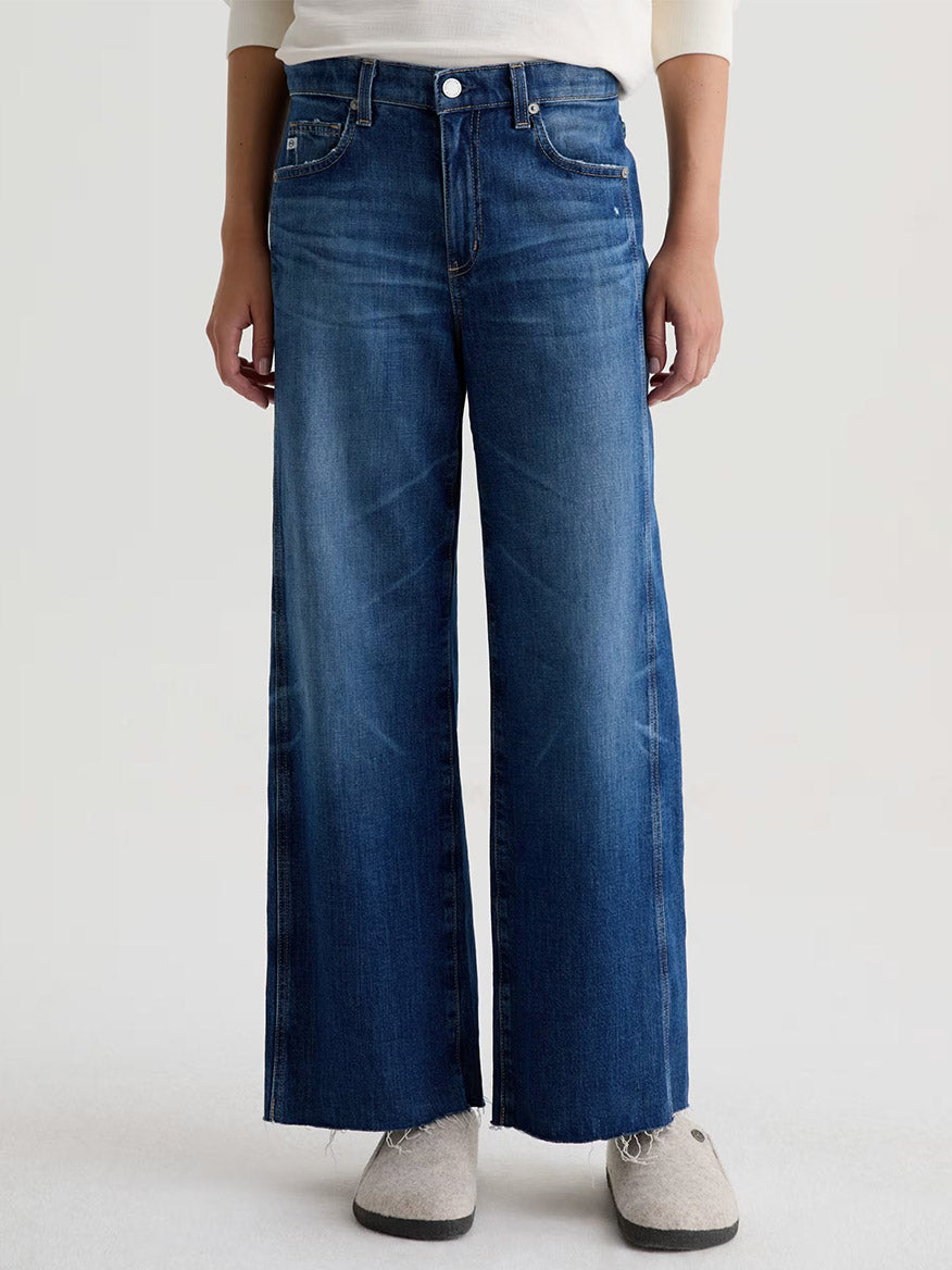 A person wearing AG Jeans Saige Wide Leg Crop in Watermist made from Candiani Mill comfort stretch denim with a raw hem and light gray shoes, standing against a plain background.