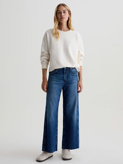 A person faces forward wearing a loose white sweatshirt, AG Jeans Saige Wide Leg Crop in Watermist, and light-colored slip-on shoes against a plain background.
