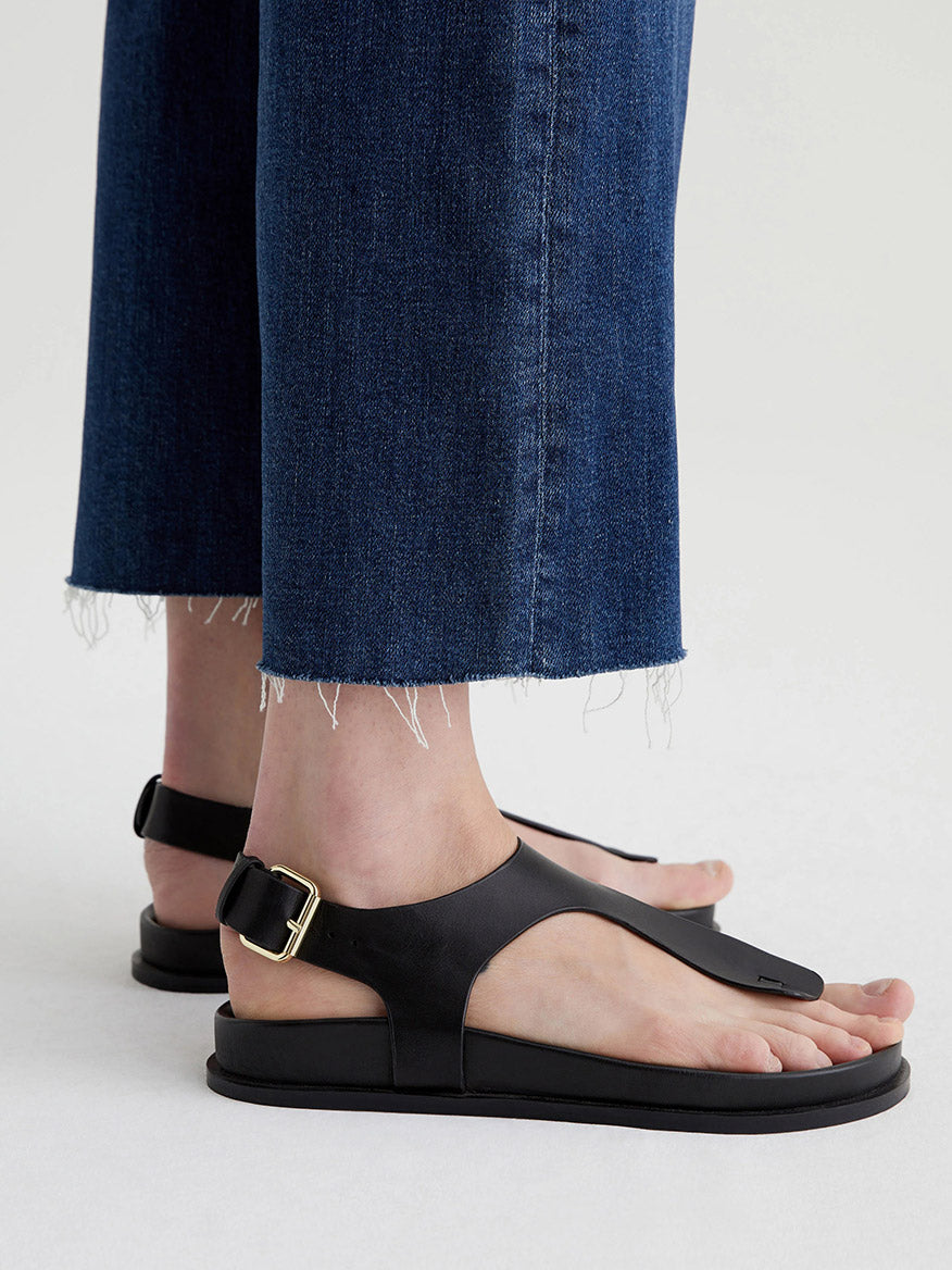 Close-up of a person wearing AG Jeans Saige Wide Leg Crop in Plume, featuring frayed hems and a dark blue wash. They pair the jeans with black leather sandals adorned with a gold buckle, standing on a light-colored surface with their toes partially visible.