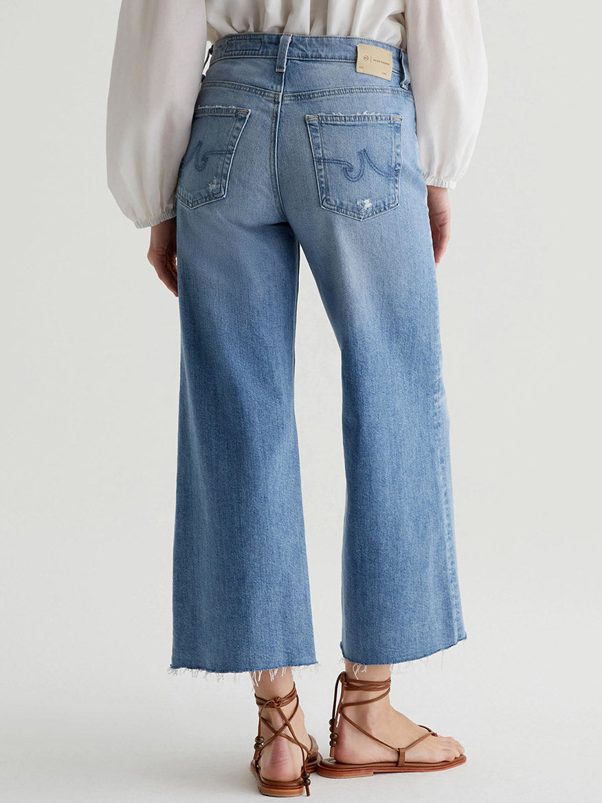 A person with a tucked-in white blouse is wearing high-waisted, light blue AG Jeans Saige Wide Leg Crop in 22 Years Palma with frayed hems and tan sandals while standing with their back facing the camera.