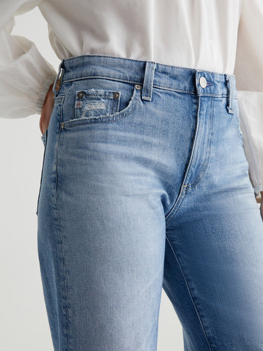 Close-up of a person wearing AG Jeans Saige Wide Leg Crop in 22 Years Palma with a high-rise waist and a white blouse. The person's right hand is partially in their jeans pocket.

