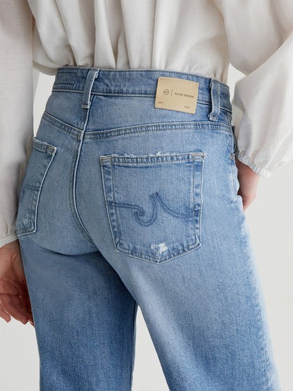 A person wearing light blue AG Jeans Saige Wide Leg Crop in 22 Years Palma with a label on the waistband and a white long-sleeve shirt. The jeans have back pockets with a curved seam design and feature cropped legs for added style.