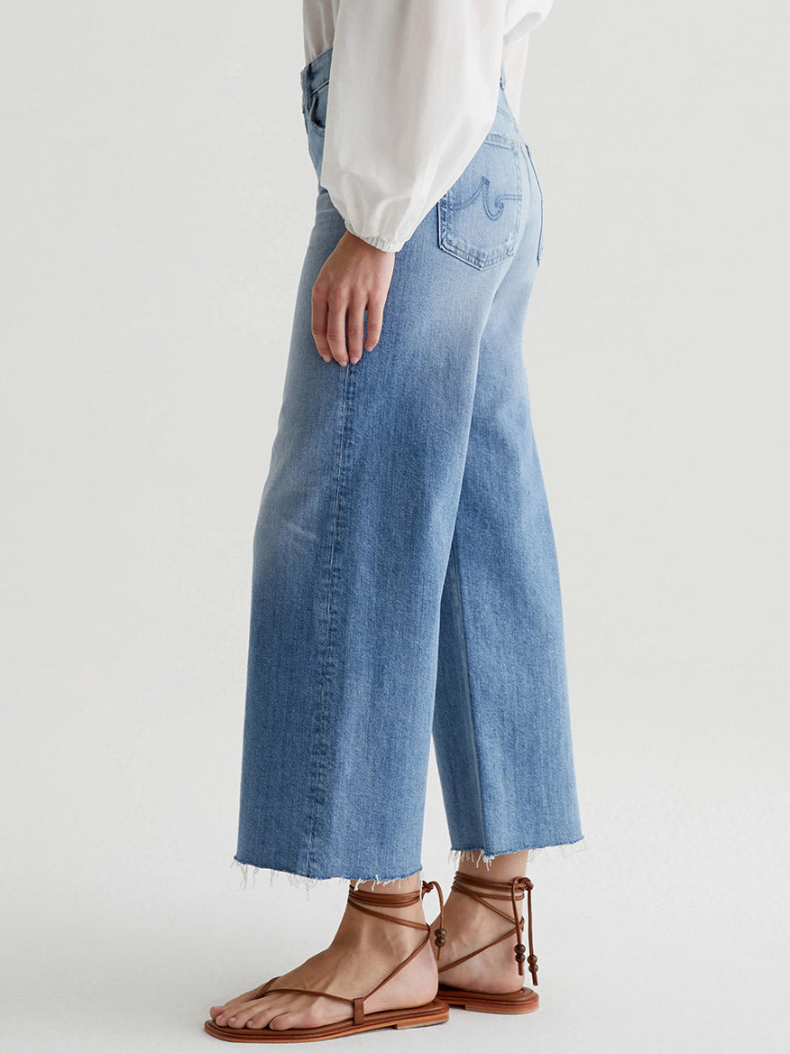 A person wearing AG Jeans Saige Wide Leg Crop in 22 Years Palma, a white long-sleeve top, and brown sandals stands against a plain background.