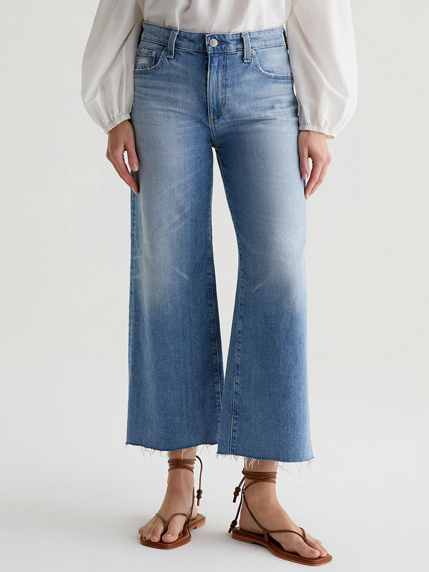 Person wearing high-waisted, wide-leg, light-wash jeans AG Jeans Saige Wide Leg Crop in 22 Years Palma with a frayed hem and a white blouse. The vintage comfort stretch denim is complemented by brown strappy sandals. The background is plain and light-colored.