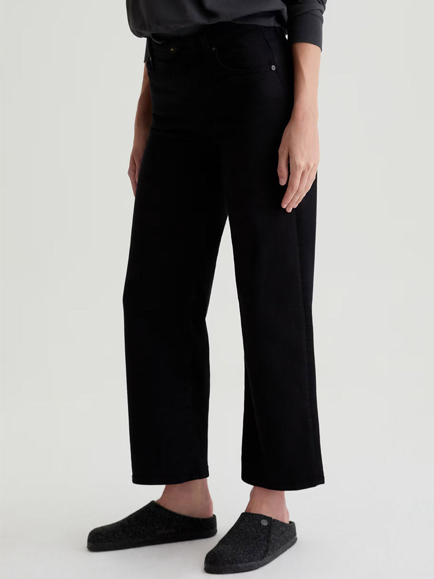 A person models AG Jeans Saige Wide Leg Crop in Super Black with dark slippers against a plain light background, highlighting the comfort stretch denim.