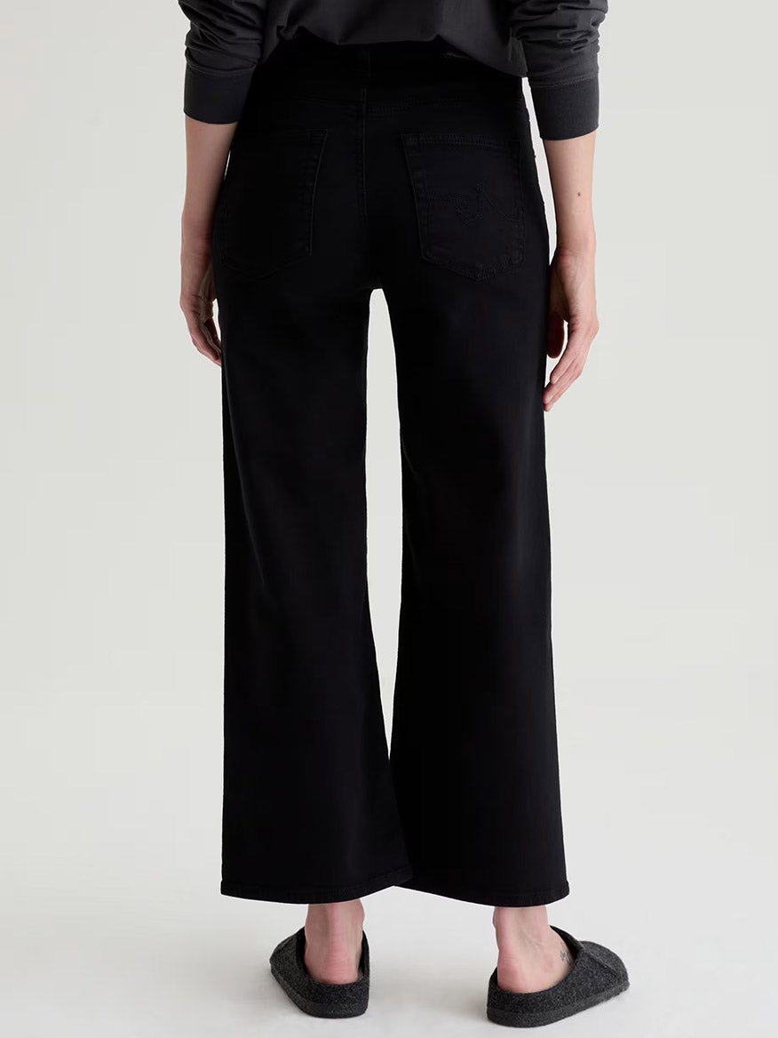 Person wearing AG Jeans Saige Wide Leg Crop in Super Black, made from comfort stretch denim, paired with dark flats, shown from the back against a plain light background.