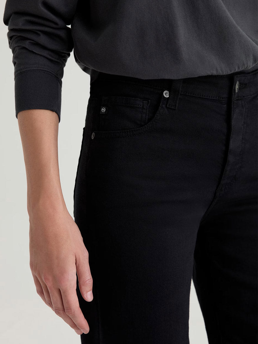 A person wearing the AG Jeans Saige Wide Leg Crop in Super Black is shown from mid-torso to thigh, with one hand relaxed at their side and paired with a long-sleeve black shirt.