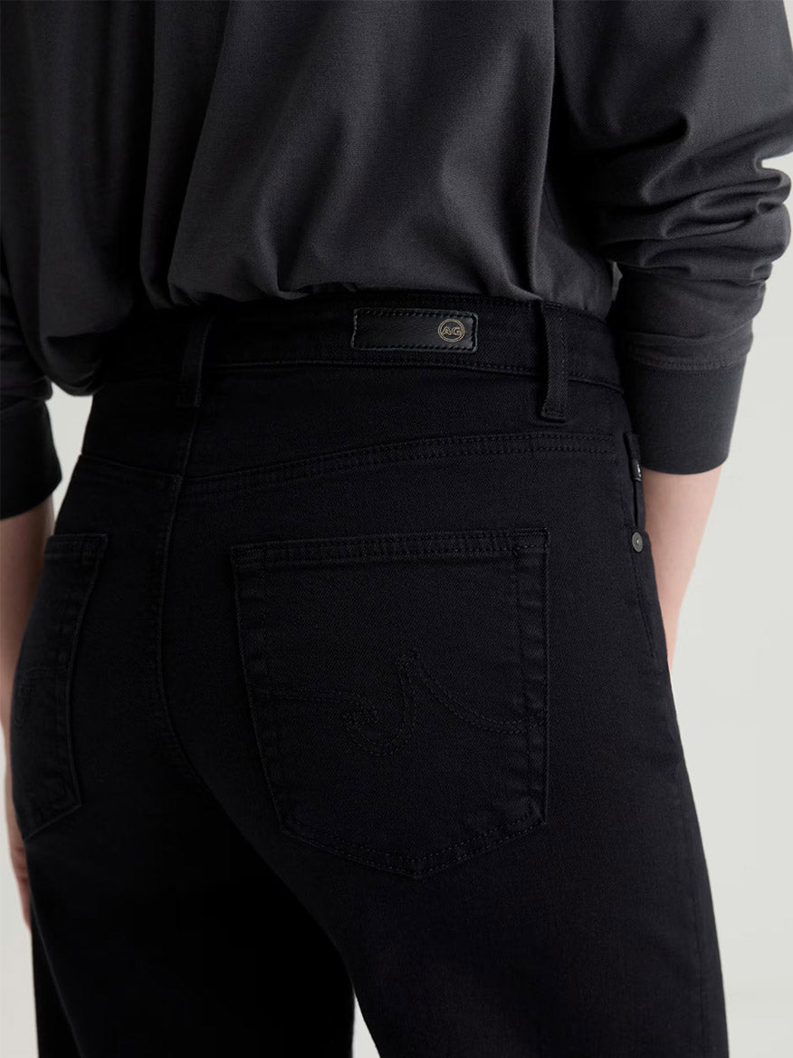 Person wearing AG Jeans Saige Wide Leg Crop in Super Black and a black long-sleeve shirt, shown from the back with focus on the jeans' pocket and waistband.