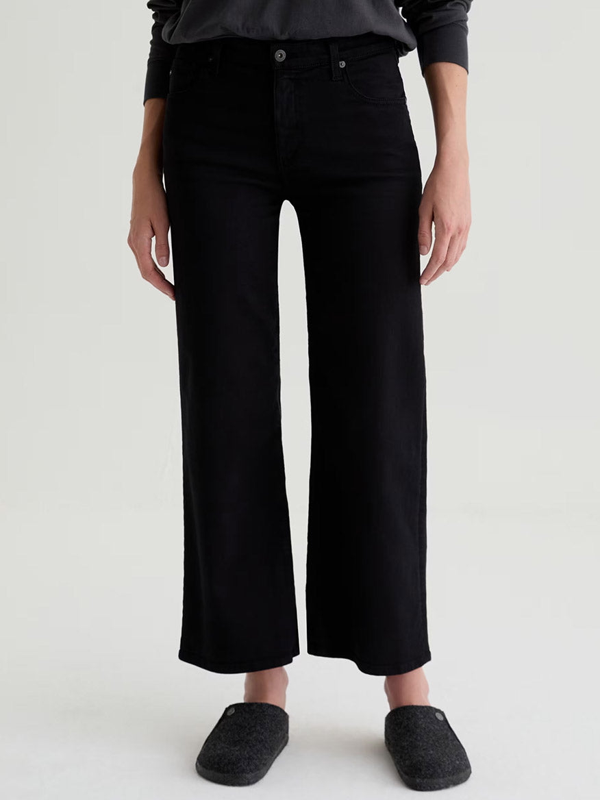 Wearing AG Jeans Saige Wide Leg Crop in Super Black, a dark long-sleeve top, and gray slippers, this person stands against a plain light background.