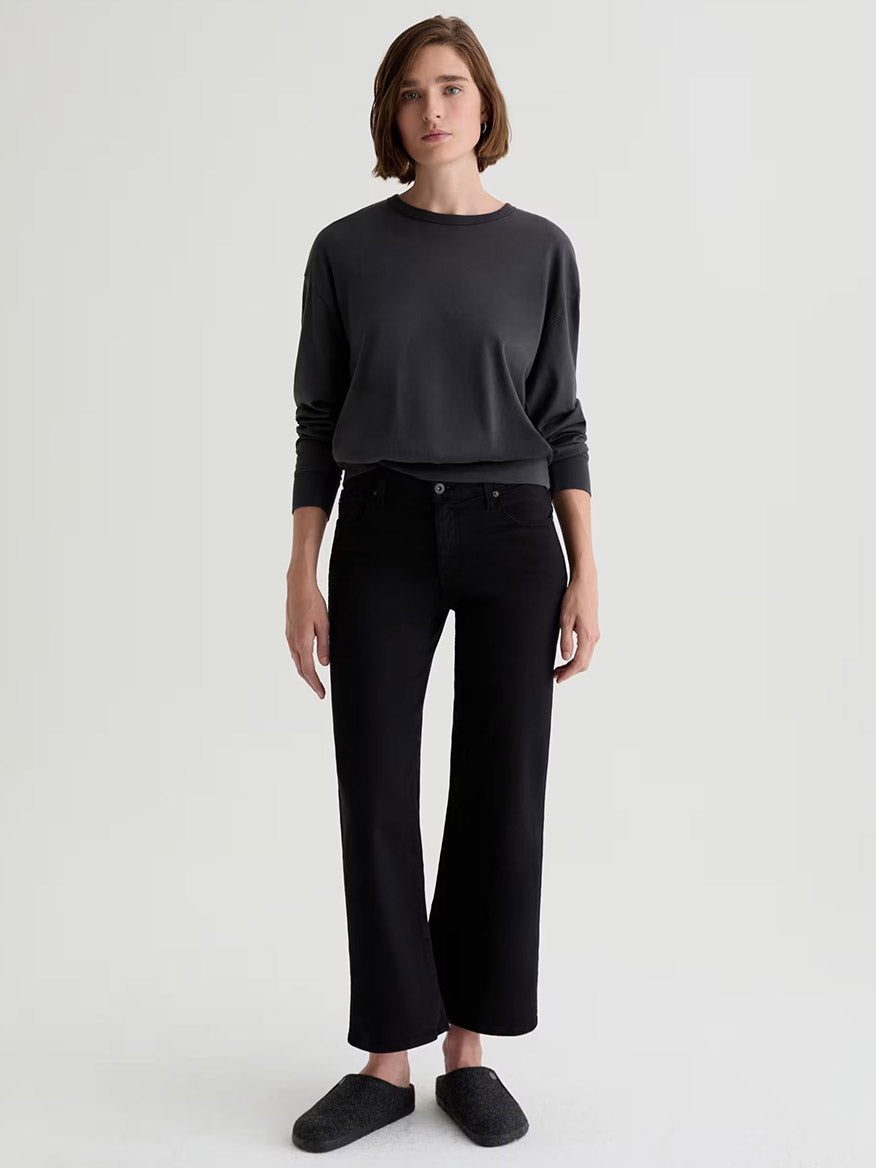 A person stands against a plain background, wearing a dark long-sleeve shirt, black pants, dark slippers, and AG Jeans Saige Wide Leg Crop in Super Black.