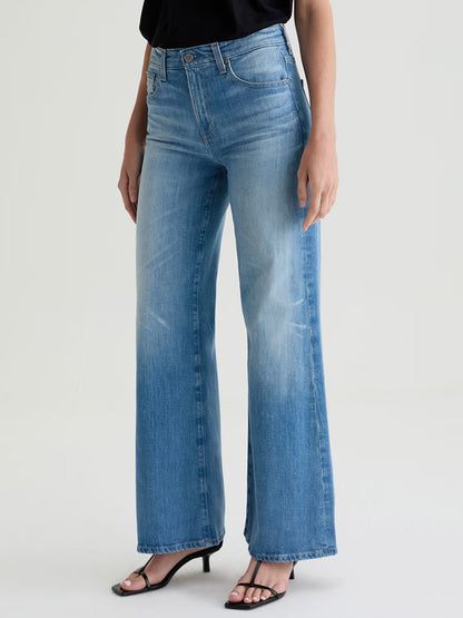 A person wearing AG Jeans Saige Wide Leg Mid-Rise in 18 Years Bisou, a black top, and black heeled sandals stands against a plain light background.