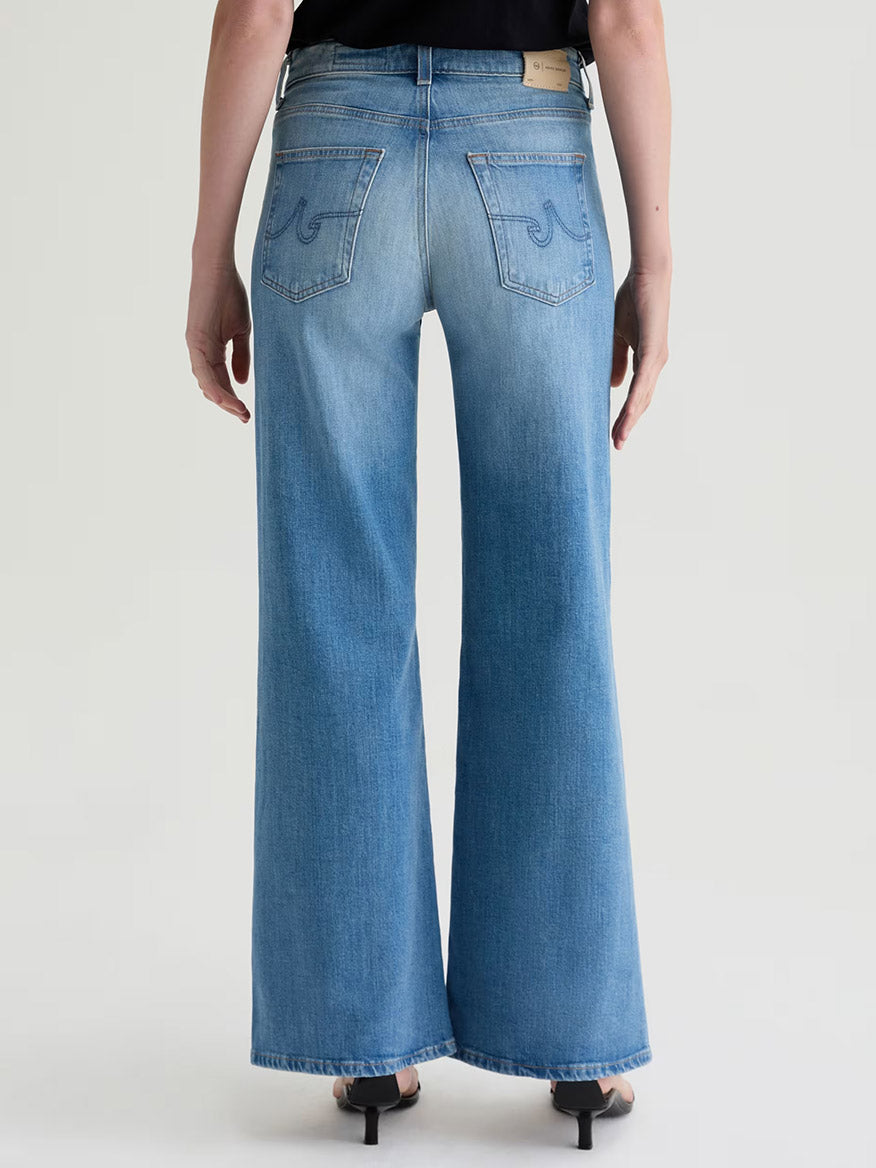 A person wearing AG Jeans Saige Wide Leg Mid-Rise in 18 Years Bisou and black heeled sandals stands with their back to the camera against a plain background.