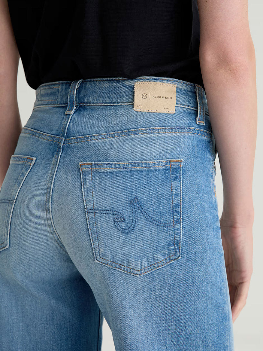 Close-up of a person wearing AG Jeans Saige Wide Leg Mid-Rise in 18 Years Bisou, paired with a black top, showcasing the back pockets and beige brand label on the waistband.