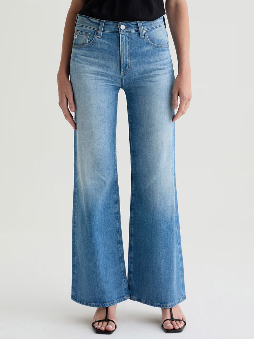 A person wearing AG Jeans Saige Wide Leg Mid-Rise in 18 Years Bisou with black heeled sandals stands against a plain background.