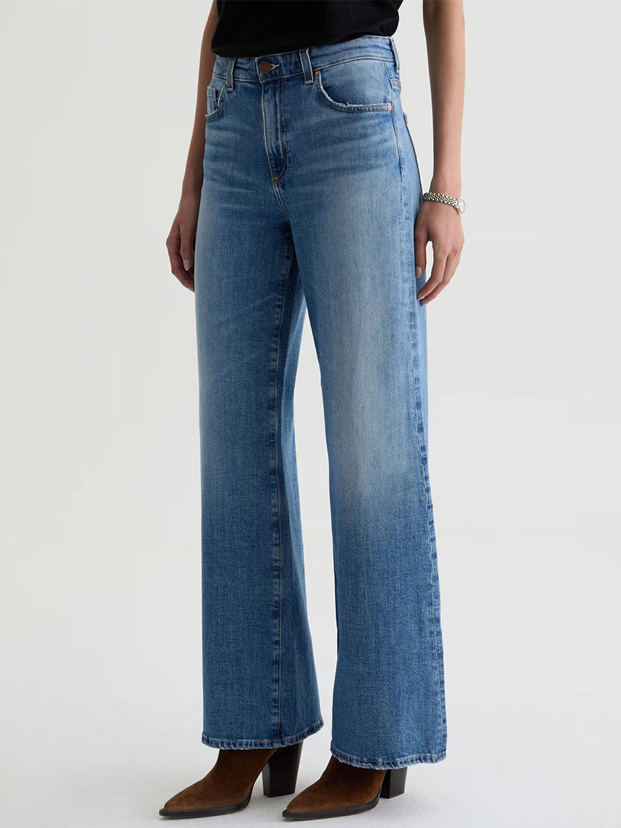 Someone stands against a plain background wearing AG Jeans Saige Wide Leg Mid-Rise in American West, a black top, and brown heeled boots.