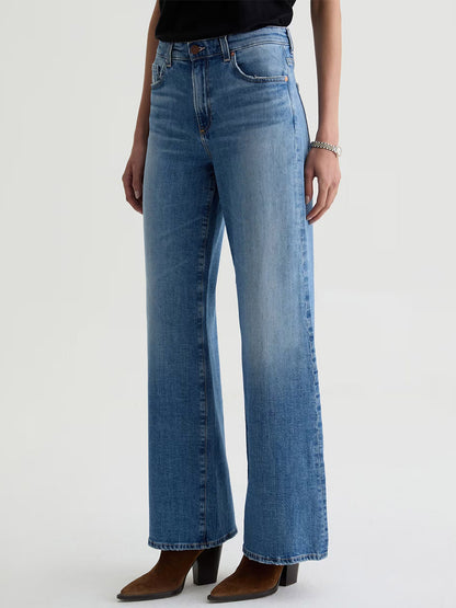 Someone stands against a plain background wearing AG Jeans Saige Wide Leg Mid-Rise in American West, a black top, and brown heeled boots.