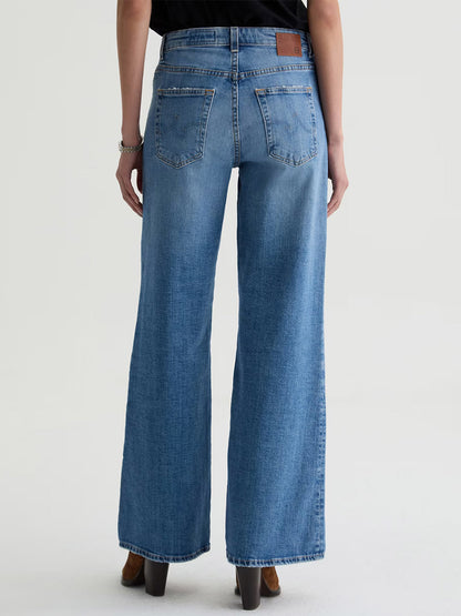 Model wearing AG Jeans Saige Wide Leg Mid-Rise in American West, shown from the back against a light backdrop. Denim features two back pockets, a brown leather waistband patch, and is made from comfort stretch fabric.