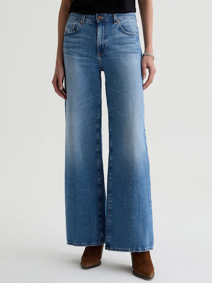 Person wearing AG Jeans Saige Wide Leg Mid-Rise in American West (light blue), a black top, brown pointed-toe shoes, and a bracelet, standing against a plain light background.