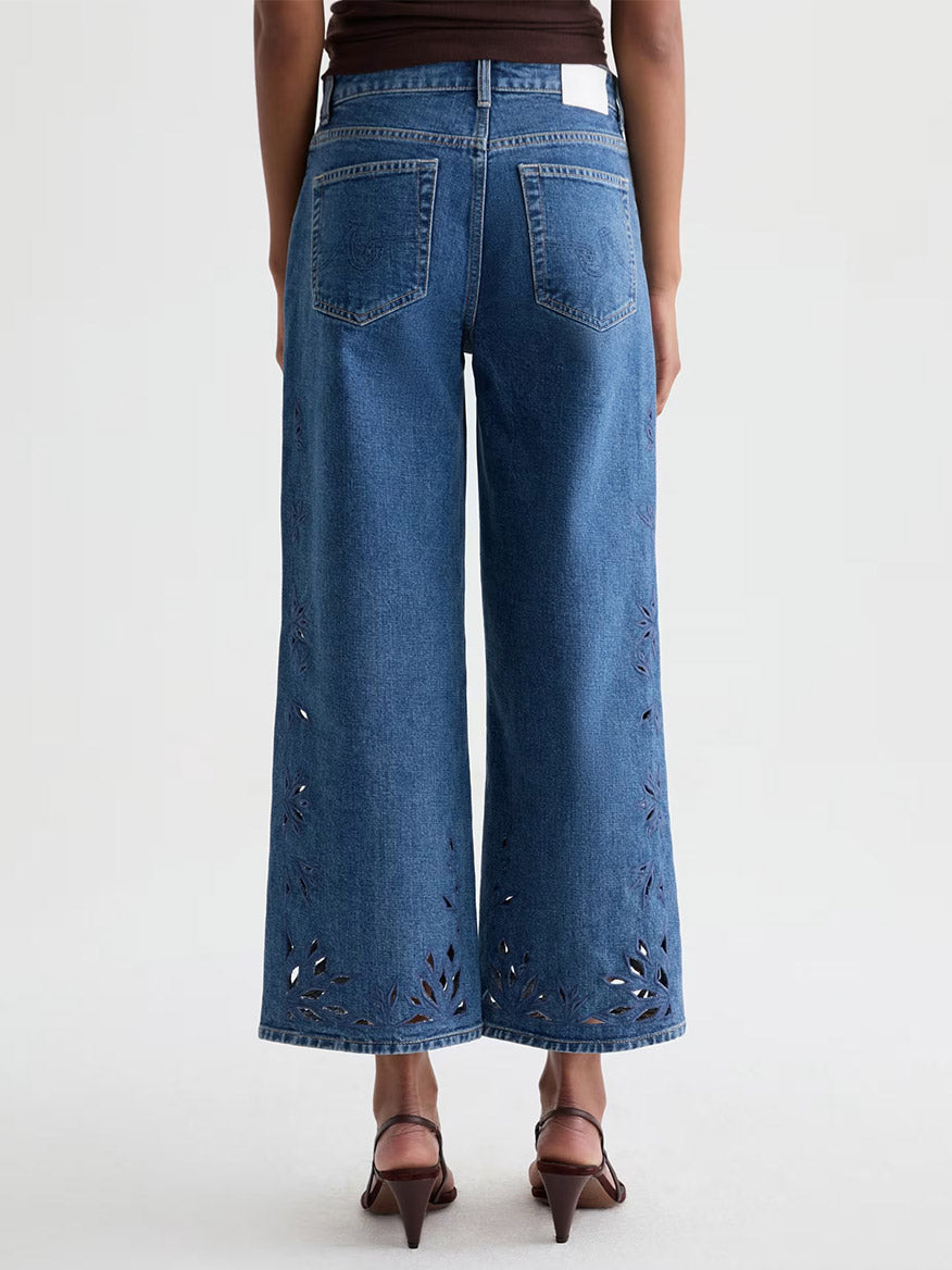 Person wearing AG Jeans Saige Wide Leg Crop in Malbec, featuring a geometric floral hem design and brown heeled sandals, shown from the back against a plain background.